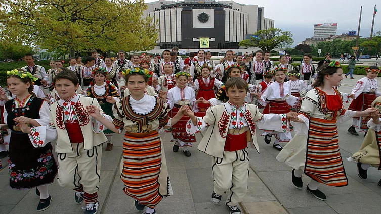 Kinder und Jugendliche in traditionellen bulgarischen Trachten tanzen gemeinsam einen Volkstanz im Freien.